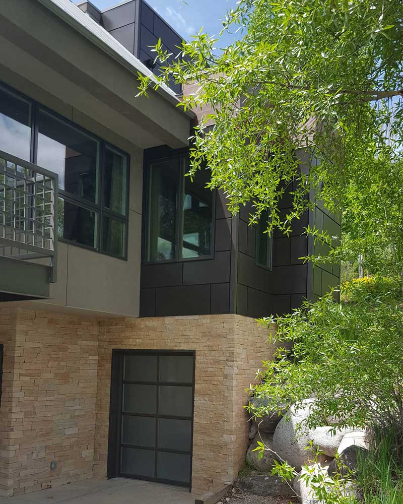 A wide shot of a transparent glass window, gray and black brick walls, and a green plant outside of a modern house design.