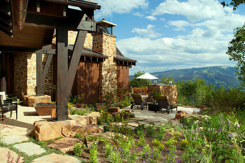 A wide shot of a house exterior on a hillside with a bench, a flower garden, and a mountain view in the background.